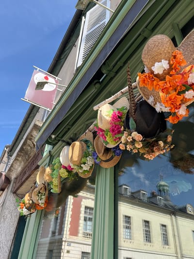 The image shows a storefront in Rambouillet, France, decorated with an assortment of hats adorned with colorful artificial flowers and feathers. The hats are displayed on a wooden beam suspended above the shop window. The storefront itself has a green frame and is large and reflective, showing a reflection of buildings across the street, including a domed structure. Above the storefront, a sign reads "L'Apiculteur". The sky is clear and blue. There are no people visible in the image.
