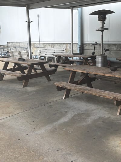 An outdoor seating area with several wooden picnic tables and benches is shown. A patio heater stands in the background, along with a small scooter. The ground is concrete, and the area is covered by a corrugated metal roof supported by metal poles.