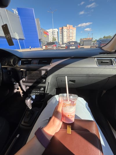 The photograph captures a view from inside a car, looking out through the windshield and side windows. In the foreground, a hand holds a plastic cup with a pink beverage and a straw, resting on a brown leather handbag. The interior of the car is visible, including the dashboard with the radio and air vents. Outside, the car is in a parking lot or on a street with other vehicles. A large blue building with a gray facade is prominent on the left, followed by a shopping center with advertisements. Further in the background, apartment buildings and a food stall are visible under a clear blue sky with scattered clouds. The scene is consistent with an urban or suburban environment. Based on the provided information, this scene is located in Fedorino, Russia.