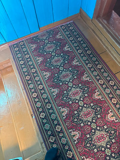 The image shows a close-up, elevated view of a long, oriental rug laid on a wooden floor. The rug features a repeating intricate pattern of red, black, white, and gold geometric and floral motifs. The floorboards are a warm, reddish-brown color and appear to be somewhat worn. In the background, to the left, is a wall constructed of bright blue wooden planks. To the right, a dark brown wooden doorframe is visible. The lighting suggests an indoor environment, possibly a hallway or entryway. There is a dark blue sock visible on a person's foot at the bottom right of the frame. The location is specified as Чуй-Атасево, Russia.