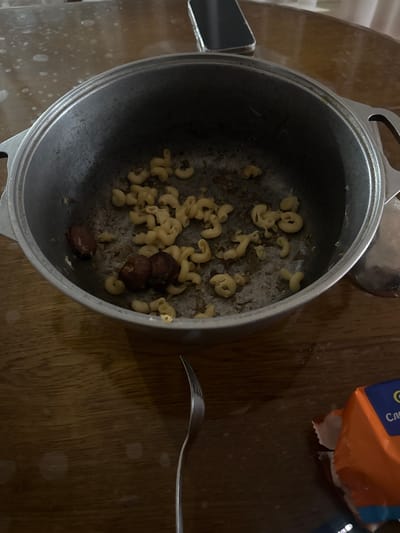 This media file displays a close-up, overhead shot of a pot containing leftover macaroni and three pieces of what appear to be small, dark sausages or olives. The pot is made of a dull, metallic material and rests on a wooden surface. Several pieces of macaroni are scattered inside the pot, some of them appearing slightly dry. To the right of the pot, a silver fork is positioned, its tines resting on the wooden surface. In the upper right corner of the frame, a portion of a food package with orange and blue accents is visible. Some of the text on the package is cut off, but the word "Cro" is discernible. The lighting suggests an indoor setting, possibly during the day or evening, with some subtle light reflections on the wooden table. There are no people visible in the image. The scene appears to be in a home kitchen or dining area.