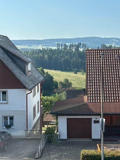 The image captures a view of the German Black Forest, with a focus on the village of St. Märgen. In the foreground, two houses dominate the scene. The house on the left features a white and gray facade with a prominent dark gray roof. Its windows have white frames, and one window on the lower floor contains a small potted plant. A dark-colored car is parked in the gravel driveway. The house on the right has a brown tiled roof and a white garage door with a number "14" visible above it. A tall, slender light post stands in front of this house.
Behind the houses, the landscape unfolds with rolling hills and dense evergreen forests. The sunlit slopes of the hills are covered in green grass, dotted with trees. The sky is clear and blue, indicative of fair weather. In the far distance, a range of hazy blue mountains forms the horizon. The overall impression is a tranquil, rural setting bathed in bright daylight.