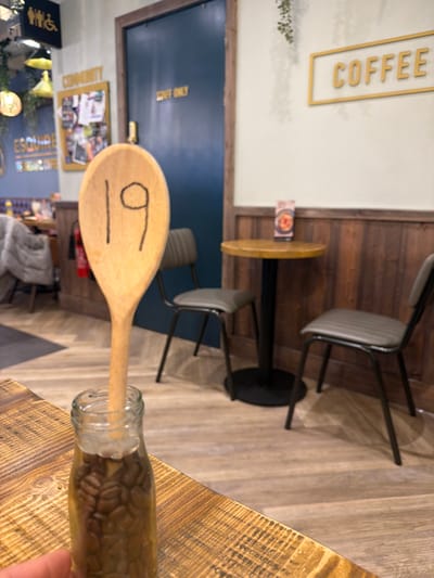 The image shows an interior space, possibly a cafe or coffee shop, with a wooden spoon inserted into a bottle filled with coffee beans resting on a wooden table. The spoon has a drawn face on it. Several chairs are positioned around small tables with a wooden countertop. In the background, there's a door with "STAFF ONLY" written on it, along with a corkboard displaying various images and announcements. A "COFFEE" sign is visible on the wall. The overall scene is rotated, so details are viewed from an unusual angle. The exact location in Mole Valley, United Kingdom, is not discernible from the image.