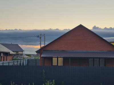 This media file shows a residential scene in Iskra, Russia, during the evening. The sky is filled with soft, warm hues of orange and yellow near the horizon, transitioning into a pale blue as it rises. A dark, elongated cloud bank stretches across the middle ground, with smaller, scattered clouds above.
In the foreground, a dark, textured wooden fence runs horizontally across the image, with some sparse green vegetation and purple wildflowers growing along its base. Behind the fence, a property with a dark metal fence is visible, separating it from neighboring houses.
The main subject is a house constructed with red brick, featuring a steeply pitched, dark metal roof. A covered porch with a similar dark metal roof extends from the front of the house, with two tall windows visible behind it. Another house with a lighter colored metal roof and brick accents is seen to the left, partially obscured by the red brick house.
In the distance, more houses with various roof styles and colors can be seen, along with power lines and utility poles, suggesting a suburban or rural residential area. The overall atmosphere is calm and serene, indicative of the end of the day. There is no visible text in the image.
