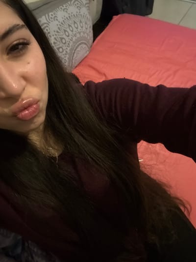 A close-up, high-angle selfie depicts a female individual indoors. The specific location in the city of Arthès, France, cannot be determined from the visual evidence. The individual, positioned slightly off-center to the left, has long, dark brown hair covering most of her left shoulder and chest. She wears a dark burgundy long-sleeved top and a thin metallic chain necklace is faintly visible. Her head is tilted, and her lips are puckered in a "duck face" expression, covered with a clear, reflective lip gloss. Her left eye is partially visible, showing dark eyeliner and what appears to be false eyelashes.
In the background, to the right and behind the individual, a bed or soft furnishing is covered with a coral-pink sheet or blanket. To the left, behind her head, a grey pillow with a white, circular, patterned design is visible. A dark object, possibly an item of clothing or a bag, is faintly discernible in the upper right background. The lighting suggests an indoor setting with artificial light sources.