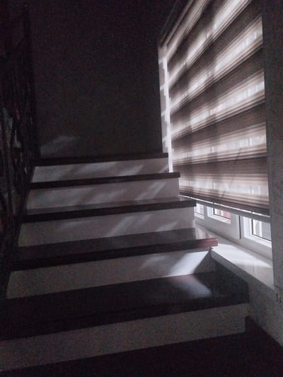 The image shows a modern staircase with dark wooden treads and white risers. The staircase is illuminated by light filtering through a striped roller blind in a window to the right. The overall scene is dimly lit, with shadows cast by the blinds on the stairs. The location in Mady, Kyrgyzstan, is not discernible from the image itself.