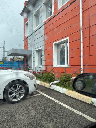 The image depicts a section of a building with an orange and grey facade, located in Elektrogorsk, Russia. The building has several windows and a downspout running vertically along its side. In front of the building, there are rose bushes with pink flowers. A white car is parked in a parking spot marked with white lines on asphalt. The scene appears to be overcast, with rain evident on the car's windshield and the building's facade. In the background, there are electrical towers and a blue fence. A car's side mirror is visible in the lower right corner of the image.