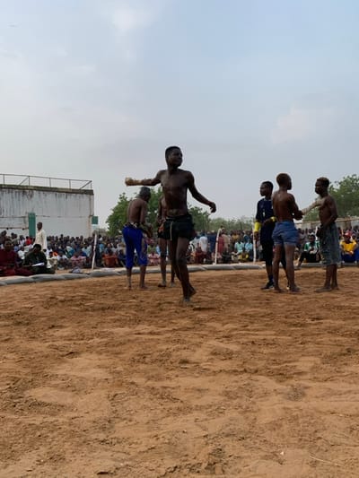 The image shows a wrestling match taking place in Sokoto, Nigeria. Five men are in the center of a sandy wrestling ring, with one man in the foreground, his back to the camera. He is shirtless, wearing brown shorts and a blue sash. He is facing two men, one of whom is in a fighting stance. The other is shirtless, wearing blue shorts, and has his back to the camera. The other two men are shirtless, with one wearing dark blue shorts and the other wearing dark blue pants. All are in a wrestling stance. There are a large number of people in the background, watching the match. They are sitting or standing along the edge of the ring. There are trees and buildings in the distance. The ground is sandy. The sky is cloudy. The location of the wrestling match is 13.049 degrees latitude and 5.238 degrees longitude, which is in the city of Sokoto, Nigeria.