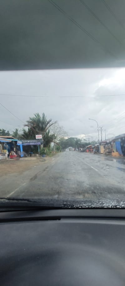 The image captures a road scene from the perspective of a vehicle's interior, looking out through a wet windshield. The weather appears overcast and grey, suggesting a recent rain or impending shower, with raindrops visible on the glass.
The asphalt road, visibly damp or wet, stretches into the distance, appearing to be a two-lane path with a white line marking the right edge in the midground. On the left side of the road, several small structures resembling shops or stalls are visible, some with blue awnings. Lush green trees, including palm fronds, stand behind these structures. A sign with red lettering reads "GELUMPANG", and below it, a blue sign displays "GAPU KRIPIK GELUMPANG", indicating a snack or chip shop.
To the right side of the road, more small buildings or stalls with varied colored tarps (red, yellow, blue) line the roadside. A utility pole with power lines and a street light fixture are also present. In the far distance, the road appears to pass under an archway formed by trees, with a vehicle, possibly a truck, barely discernible beyond. The foreground shows the car's dashboard and the lower part of the windshield, emphasizing the indoor perspective. The scene suggests a rural or semi-urban area in Glee Gogo, Indonesia, likely during daytime given the ambient light, despite the heavy clouds. No people are clearly visible engaged in specific activities, though the presence of shops implies commercial activity.