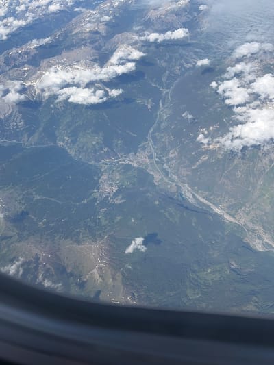 This detailed aerial image, captured from an aircraft window with a partial view of the window frame visible at the bottom, depicts a vast mountainous landscape in the region of Usseaux, Italy, and its surroundings. The scene is dominated by rugged, snow-capped peaks in the upper left, transitioning into densely forested slopes and deep valleys.

A prominent, wide valley traverses the central and right portions of the image, featuring a winding river, likely the Dora Riparia or a tributary, which appears as a light grey ribbon. Parallel to the river, a major multi-lane highway or autostrada with associated infrastructure, including bridges and interchanges, cuts through the valley floor. Several settlements and towns, characterized by clusters of small, light-colored buildings, are visible along the riverbanks and highway, extending up the lower slopes of the mountains.

The majority of the non-rocky terrain is covered by dense, dark green coniferous forests, interspersed with lighter green clearings or meadows on gentler slopes. Scattered patches of residual snow are visible on the higher mountain peaks and some upper slopes, suggesting a late spring or early summer timeframe. Numerous white, fluffy cumulus clouds are dispersed across the sky, casting distinct shadows upon the mountainsides and forested areas below. The overall visibility is clear, allowing for sharp detail of the terrain, infrastructure, and atmospheric elements. No people or animals are discernible at this altitude.
