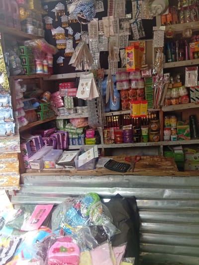 This image captures the interior of a retail stall or small shop in Birnin Kudu, Nigeria. No individuals are visible.

The scene features multiple wooden shelves heavily laden with merchandise. The upper shelves, particularly on the right, display numerous pieces of costume jewelry, including earrings and pendants, individually packaged on small cards and arranged against a dark background. Many of these items are gold-toned. Above this display, a blue and white striped fabric item is visible.

The shelves below present a dense array of packaged goods. These include:
- Small, round plastic containers, some white, some with pink lids, likely cosmetics or creams.
- Rectangular boxes, some green, some red and white, with branding such as "MYMY" and images of women.
- Various bottles and jars containing liquids or creams, in colors like blue, green, and amber.
- Stacked boxes, some purple, others featuring different designs.
- Clear plastic containers holding small items.
- Larger jars containing what appears to be honey or oil, along with other cylindrical containers in various colors (gold, purple, green).

A wooden counter is present in the lower-mid section of the image. On this counter, a black electronic calculator is positioned to the left, alongside a pair of scissors with red handles. The wall behind and below the counter consists of corrugated metal sheeting, indicating a basic construction.

The foreground contains various items primarily wrapped in clear plastic bags, suggesting soft goods or small accessories. These include what appear to be baby hats or garments, one pink with multi-colored letters spelling "S.D.O.G.S." Other packaged items, possibly tissues or snacks, with visible branding like "Duro" and "Sandal," are partially visible on the far left.