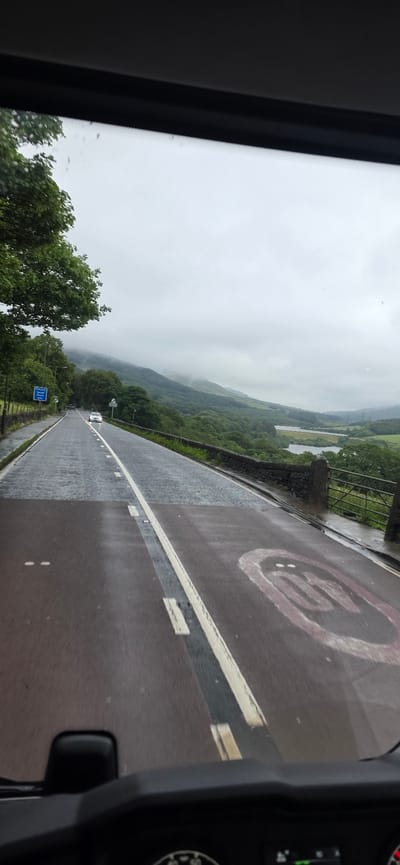 A wet asphalt road curves to the right, leading into a valley with misty green hills and two bodies of water. The road has white lane markings and a faded circular speed limit sign, possibly indicating 30. A white car is visible in the distance on the road. Trees line the left side of the road, and a stone wall with a metal gate runs along the right. The sky is overcast and cloudy. The image appears to be taken from inside a vehicle, looking out through a windshield. The location is in High Peak, United Kingdom.