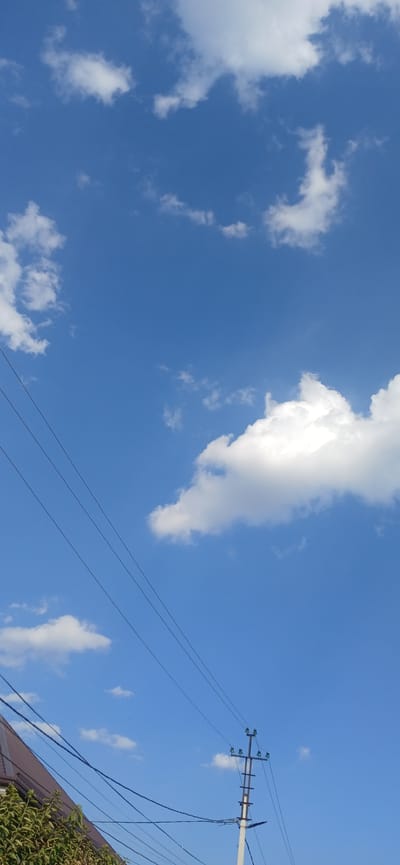 A vertical, medium shot captures a clear blue sky with scattered white cumulus clouds. In the lower left corner, a portion of a brown-tiled roof and green foliage is visible. Diagonal black power lines extend across the frame from the bottom left. To the right of the power lines, a utility pole with multiple crossbars and insulators stands, supporting more power lines. The scene appears to be outdoors in an urban or suburban environment, potentially in Daliguzar, Uzbekistan, given the context, under daylight conditions. No people or animals are present in the image.