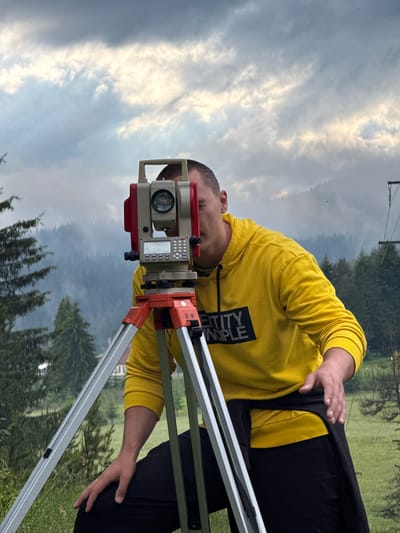 A man in a yellow hoodie with "ENTITY PRINCIPLE" printed on it is using a surveying instrument on a tripod. He is positioned in front of a backdrop of misty, tree-covered mountains. The sky above is cloudy and appears to be either dusk or a heavily overcast day. The foreground shows green grass and some trees. The overall scene suggests an outdoor surveying activity in a mountainous or hilly area, potentially Yundola, Bulgaria, given the context.