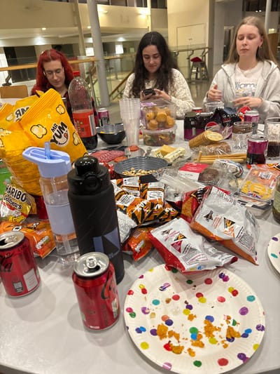 Three women gather around a table laden with snacks. One woman in red hair and glasses sits in the foreground. Another woman with dark curly hair uses her phone. The third woman, with blonde hair, sits near the top of the frame. The table is covered with an assortment of treats including chips, Haribo gummy bears, "Mr. Yum" popcorn, breadsticks, Coca-Cola cans, and cheese. A colorful, polka-dot paper plate with crumbs rests on the right side of the frame. A black water bottle lies near the table's edge.