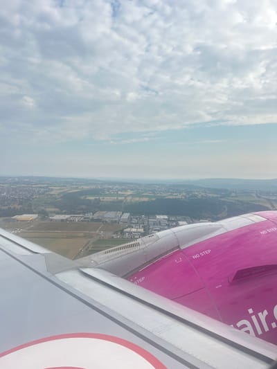 This is an aerial, eye-level view from an airplane window looking out at the landscape below. The foreground is dominated by the wing of the aircraft, a portion of which is painted bright pink with "air.com" and "NO STEP" markings visible. The land below shows a mixture of urban and rural elements. There are buildings, some appearing to be industrial facilities with large roofs and parking areas, interspersed with agricultural fields and patches of green trees. A town or city is visible in the mid-ground, with houses and streets stretching into the distance. The horizon is hazy, with rolling hills or mountains in the far distance under a sky filled with scattered clouds. The location is identified as Echterdingen, Germany.