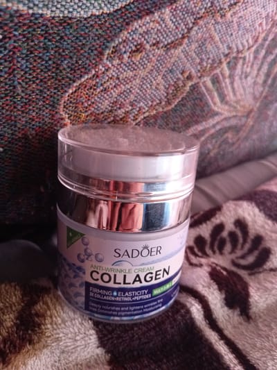 A close-up, eye-level, interior shot captures a round, plastic jar of SADOER ANTI-WRINKLE CREAM COLLAGEN. The jar is silver and white with a clear lid, and sits on a textured, patterned fabric. The background fabric is a dark, multi-colored tapestry-like material in shades of blue, red, and green, with a woven texture. The jar's label features text including "SADOER," "ANTI-WRINKLE CREAM," and "COLLAGEN," along with smaller text describing its benefits. The lighting is soft, creating subtle reflections on the jar's surface. The image is taken in Monastirische, Russia.