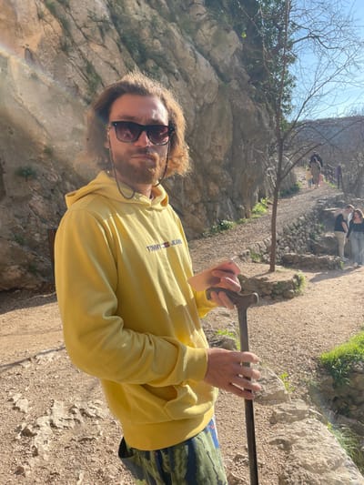 A male subject stands on a gravel path in a sunny, rocky outdoor environment in Dračevice, Bosnia and Herzegovina. The subject, with light brown hair and a beard, is centrally framed, looking directly forward. He wears dark sunglasses with a black beaded chain, a yellow hooded sweatshirt featuring the "TOMMY JEANS" logo, and camouflage-patterned trousers. His hands grip a dark walking stick; a small, light-colored object is held between the thumb and forefinger of his left hand. To the subject's left, a rocky cliff face is visible. To his right, a dirt path with stone retaining walls and sparse vegetation, including a small, bare-branched tree, ascends into the background. Several other individuals are observed walking along this path. Strong sunlight illuminates the scene, casting shadows and creating a rainbow-colored lens flare in the upper left frame.