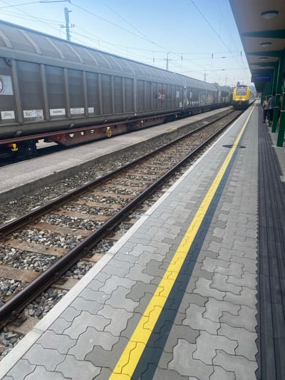 The image shows a train platform in Wulkaprodersdorf/Vulkaprodrštof, Austria. On the left, a long freight train is parked on a track. The freight cars are grey and appear to be carrying some form of cargo. The train has markings that include "EUROFIMA" and "Cargo". On the right side of the image, a yellow passenger train is approaching or stopped at the platform. Several people are visible on the platform, standing near the passenger train. The platform is paved with interlocking concrete blocks and has a yellow line marking the edge. The environment is outdoors under a clear blue sky. Overhead wires for the electric train are visible. A station roof structure extends from the right side of the frame.