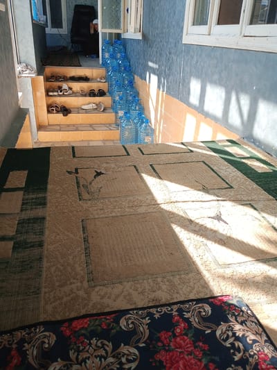 This media file depicts an indoor scene in Suzak, Kyrgyzstan, likely a home or communal space. In the background, a person wearing a white head covering and dark clothing sits in a large black chair. In front of them, a stack of blue plastic water bottles lines the steps, suggesting a supply of drinking water. The steps themselves are made of orange tiles and are lined with various shoes, including sandals and sneakers, indicating a place where shoes are removed. The foreground is dominated by a large rug with a beige and green patterned design, and another rug with a dark background and a red floral pattern is partially visible at the bottom. Sunlight streams in from windows, casting distinct shadows across the floor and walls, suggesting it is daytime. The overall impression is one of a lived-in, practical space.