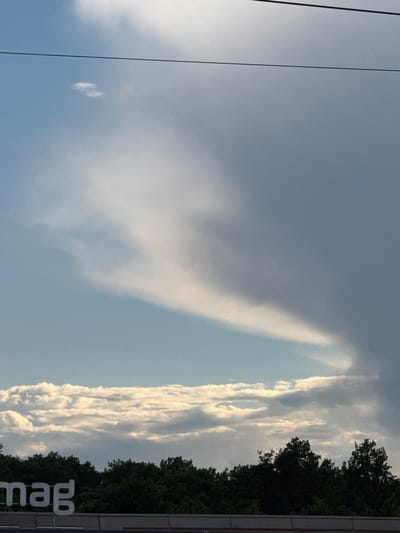 In the city of Vernier, Switzerland, this image captures a sky filled with dramatic clouds. A thin, dark wire runs horizontally across the upper portion of the frame. Below this wire, a large, wispy cloud formation stretches diagonally from the lower left to the upper right, contrasting with the clear blue sky on its left and darker, more ominous clouds on its right. Towards the bottom of the image, a dense line of dark green trees forms a solid canopy. In front of the trees, a portion of a white sign with large, blocky letters spelling "mag" is visible. The overall impression is one of a dynamic sky over a treeline, possibly near a commercial area.