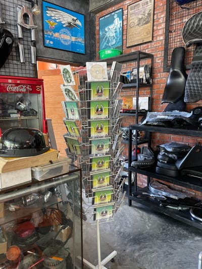 This image captures the interior of a specialized shop, presumed to be located in Ban Yid, Thailand. The environment features a combination of industrial and rustic elements, with a dark gray, gridded metal wall on the left, a dominant red exposed brick wall in the background, and a polished, dark gray concrete floor.

On the upper left wall, multiple chrome motorcycle exhaust pipes and a black motorcycle fuel tank are vertically mounted on the gridded surface. Above these components, a large, blue-framed poster displays a white graphic of an eagle, with discernible text indicating "22nd Annual Asia Pacific," associated with a motorcycle organization. Below this, a red Coca-Cola branded glass-front refrigerator is present, containing various metallic and other items.

In the lower left foreground, a glass display counter features a glossy black half-shell motorcycle helmet resting on a wooden block, alongside a clear plastic container. The interior of this display case houses an assortment of small motorcycle parts, including red and orange signal lights, wiring, and metallic components.

A white, multi-tiered metal display rack occupies the center, holding numerous media cases, specifically VCDs or DVDs. Many of these cases exhibit an eagle logo and are labeled "Phuket Bike Week 2018 Event VCD." A single optical disc is placed on the top tier of this rack.

The red brick wall in the background displays two framed posters. The poster on the left is blue, depicting two figures on motorcycles with visible text including "Mickey Rourke" and "Don Johnson." The poster on the right is sepia-toned, advertising "Wanted Bikers To Lampang Bike Night 2023" with an illustration of a motorcycle.

On the right side of the scene, a black multi-tiered metal shelving unit is fully stocked. It primarily holds various black motorcycle seats in different designs, including smooth, quilted, and studded styles, some of which are still enclosed in clear plastic packaging. Additional motorcycle components, such as covers and smaller parts, are also visible on these shelves. To the far right, another section of the dark gray gridded metal wall is visible, with more motorcycle seats or accessories hanging from it.

No individuals are present in the image, and no active interactions or actions are depicted. The scene is static, showcasing a retail or display space dedicated to motorcycle parts and memorabilia.