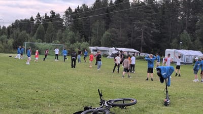 The image shows a group of children and young people playing soccer on a grassy field. In the background, there are trees, campers, and a soccer goal. A bicycle and an electric scooter are lying on the ground in the foreground. The scene appears to be a recreational area, possibly a campground or park, located in Östersund, Sweden. The time of day appears to be late afternoon or early evening, as indicated by the lighting conditions.