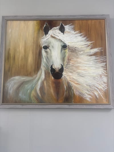 A framed oil painting of a white horse with its mane blowing in the wind is displayed on a light gray wall. The painting is horizontally oriented and features the horse's head and neck. The background is a warm, golden-brown color. The frame is a light gray, distressed wood. The painting is hanging on a plain wall in what appears to be an indoor setting. There is no information available regarding its location in the city of Pereiaslavske, Ukraine.