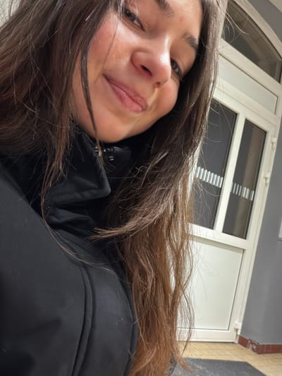 A young woman with brown hair smiles slightly, her face angled towards the viewer. She is wearing a black jacket. The background consists of a white door or wall with rectangular window panes. The lighting appears to be indoors, possibly fluorescent. The angle suggests the photo was taken from a slightly elevated perspective, looking down at the subject.