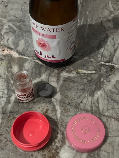 The scene depicts five items arranged on a light grey and white veined marble surface, presumably within Ksar-el-Kebir, Morocco. In the upper portion, a dark amber glass bottle labeled "ROSE WATER Distilled" and "مقطر الورد" (Muqattar Al Ward) with a 200 ml capacity is positioned upright. Below and to the left of the bottle, a small, clear glass jar contains a quantity of reddish-pink powder; its white label partially reads "AKAR EL FASSI" (أكار الفاسي). Adjacent to this jar, a small, dark grey, circular plastic cap rests upside down. In the lower portion of the image, an empty, open, circular, red plastic container base is visible. To its right, a pink plastic lid, also circular, features gold embossed text reading "VASELINA NEUTRA PERFUMADA" (Neutral Perfumed Vaseline) encircling a central graphic. No individuals are present.