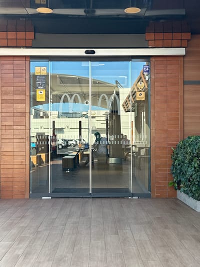 The image shows the exterior of a McDonald's restaurant in Sedaví, Spain. The entrance features automatic glass sliding doors framed by brick and wood paneling. Inside, the restaurant's iconic golden arches are visible on the glass. Several people are seen inside, including one individual wearing a green head covering who appears to be an employee. Outside the entrance, there is a wooden deck and some greenery. The overall scene suggests a typical day at a fast-food establishment.