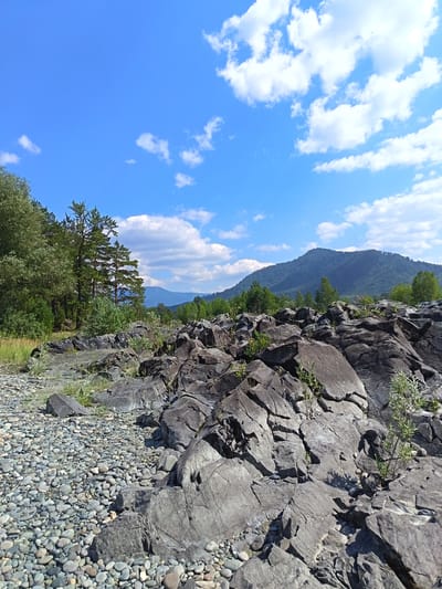 This image depicts a rocky riverside scene under a bright blue sky with scattered white clouds. The foreground is dominated by a large expanse of grey, weathered rocks, some of which are large and jagged, while others are smaller and more rounded, resembling pebbles. Sparse green vegetation grows among the rocks. To the left, a shoreline covered in smooth, grey pebbles leads towards the water. In the middle ground, a dense line of green trees, including tall pines, forms a natural barrier. Beyond the trees, rolling, tree-covered mountains recede into the distance. The sky is a vibrant blue, with fluffy white clouds providing a striking contrast. The scene is set in a natural, undeveloped environment, likely a park or natural reserve, with no human figures or man-made structures visible, except for what appears to be a small white vehicle parked in the far distance, partially obscured by trees. The location is specified as Nizhnekamcha, Russia.