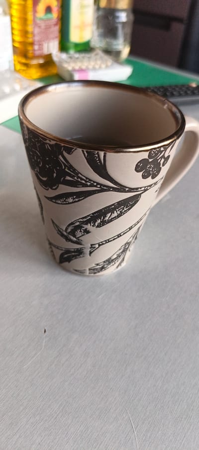 A close-up shot focuses on a cream-colored mug with a black floral pattern and a gold rim. The mug is positioned on a light-colored, textured surface that appears to be a table. In the background, out of focus, are various items including a bottle of yellow liquid, a green bottle, and what appear to be glass containers. The scene is indoors and lacks specific markers of Tikhvin, Russia, or any discernible urban environment. The image primarily showcases the mug.