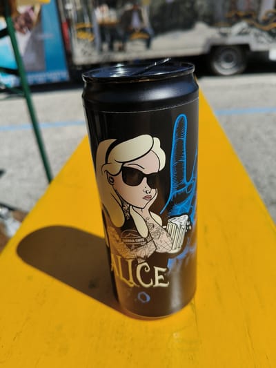 A black can with a cartoon image of a girl with blonde hair, sunglasses, and tattoos is sitting on a yellow table. The word "ALICE" is printed on the bottom of the can. A truck is visible in the background. The location is likely in Bellaria-Igea Marina, Italy, given the presence of "SARROK CAFFE" on the can, which is a local establishment.