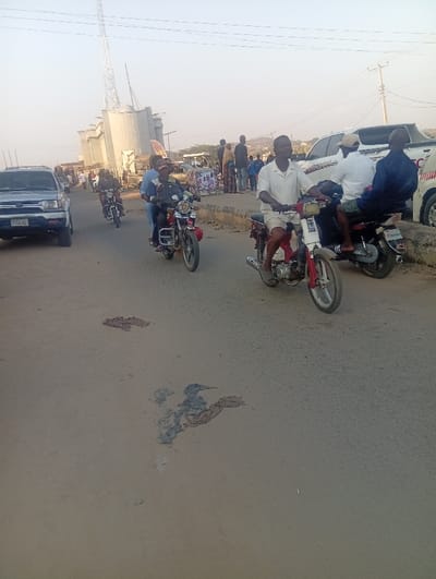 This image captures a daytime scene at Itakpe Junction, Nigeria, under clear skies. The primary subjects are several people traveling on motorcycles on a paved road.

In the foreground, a man wearing a white shirt and shorts rides a white and red motorcycle, looking forward. Behind him, two men are on a red motorcycle, with the driver wearing a dark top. Further back on the left, another individual rides a dark motorcycle, passing a silver SUV, which has a partially visible license plate reading "FGH 532 KG". To the right, two men are on a dark scooter or moped; the driver is in a light shirt, and the passenger, also in a light top, is seated side-saddle.

The background features a blend of industrial and commercial structures. On the left, large industrial silos and associated buildings are prominent, with a tall communication tower extending into the sky above them. Further down the road, additional people are visible standing or walking on the roadside, and a white pickup truck, possibly a Toyota Hilux with some red branding, is parked or driving. The road surface in the immediate foreground appears worn, showing patches of discoloration or debris.

The scene depicts a typical daily activity, with individuals commuting or traveling by motorcycle, a common mode of transport in the region. The lighting suggests it is bright and sunny.