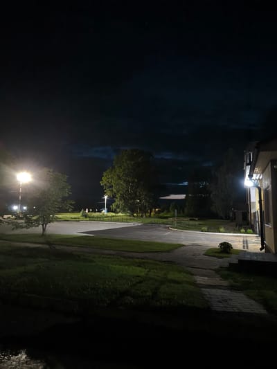 This is a vertical, eye-level, medium shot taken at night in a rural area, possibly the village of Babinichi, Babinichi rural council, Belarus. A bright street lamp illuminates a small tree in the left foreground, casting a strong shadow. To the right, the corner of a light-colored building is visible, with another light fixture attached to its side. The scene depicts a paved pathway leading to the building and a road or driveway. Beyond the immediate foreground, a green lawn is visible, and further in the distance, trees and a dark, cloudy sky are present. There are no people or animals discernible in the image. The overall atmosphere is one of quiet darkness, punctuated by artificial lighting.