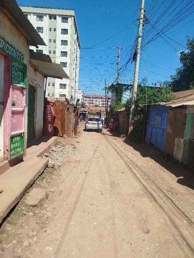 The image depicts an unpaved dirt road within Kihara ward, Kenya, under a clear blue sky. On the left, a single-story building with a pink facade and green trim displays signs for "D'S COSMETIC" listing "PERFUME, REFILL, DIAPERS, TISSUES, SANITARY," and another sign stating "SODA BARIDI WELCOME." A concrete curb runs along this structure. Adjacent to it are structures built from corrugated metal, exhibiting rust and an "airtel" banner. On the right, further corrugated metal structures are visible, painted in colors such as blue, green, and brown, with overgrown vegetation behind them. Multiple utility poles carrying numerous black wires traverse the scene overhead and along the right side. A silver compact car with an unreadable license plate is positioned centrally on the dirt road, facing away from the viewer. Indistinct human figures are visible further down the road past the car. In the far background, a prominent multi-story building with a light-colored facade and multiple windows stands on the left, while other buildings with reddish roofs are discernible in the center-right background. The scene is brightly illuminated by direct sunlight, casting shadows.