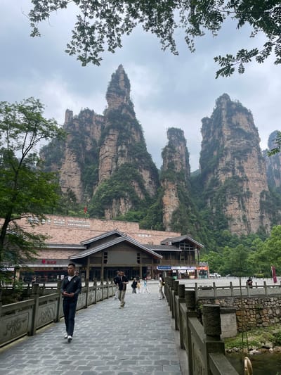 The image shows a scene in Xiehe, China. Towering rock formations with lush vegetation cover the background, creating a dramatic natural backdrop. A paved stone pathway runs horizontally across the lower portion of the image, lined by an ornate railing. A man in a dark suit stands on the pathway, looking toward the camera. Adjacent to the path is a multi-story building with traditional Chinese architectural elements. Several other people are walking on the pathway in the distance. The weather appears overcast.
