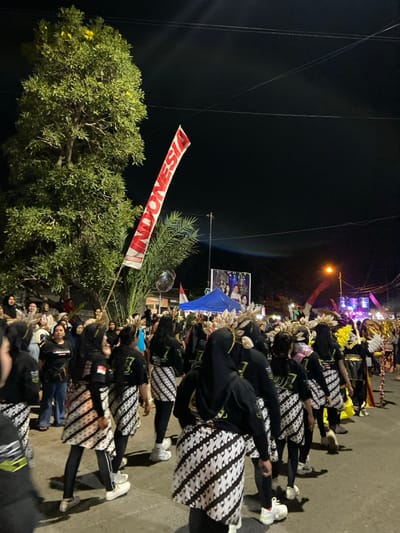 A night-time cultural parade or procession is depicted on a city street in Kedungbanteng, Indonesia. A line of individuals, primarily female, moves away from the viewer in the foreground and mid-ground. Participants wear black long-sleeved shirts, black leggings, and white-and-black patterned wrap-around skirts, consistent with a batik style. Many wear black headscarves. Elaborate feathered or decorative headpieces and back accessories resembling wings adorn several individuals, indicative of ceremonial or performance costumes. All wear white athletic shoes.

To the left, a large, leafy green tree partially conceals a crowd of spectators. A red and white banner with "INDONESIA" printed vertically in white capital letters is prominently displayed mid-left on a pole. A smaller Indonesian flag is also visible further back. A blue canopy tent stands in the mid-background, with a large illuminated screen or billboard featuring images of people behind it. To the right, a brightly lit stage or performance area with colorful spotlights (pink, purple) and sound equipment is visible, illuminated by an orange streetlamp. Overhead utility lines are present against the dark night sky.