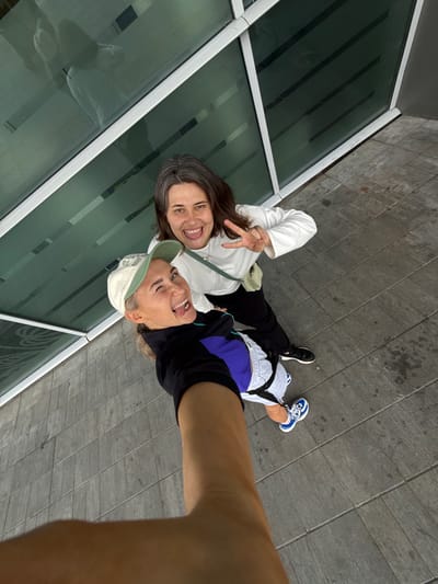 Two individuals are posing for a selfie outdoors. The person in the foreground, wearing a black jacket, purple and blue accents, light grey shorts, and a light-colored baseball cap, is sticking their tongue out at the camera. The second person, standing behind and slightly to the right, is wearing a white long-sleeved shirt, black pants, and is making a peace sign with their right hand. They are both smiling. The background consists of a modern building with large, dark green windows and a light-colored frame, and a paved walkway made of grey rectangular tiles. The overall scene suggests a casual, friendly interaction in an urban or institutional setting. There is no visual information in the image to suggest the location is Bol'shie Kabany, Russia.
