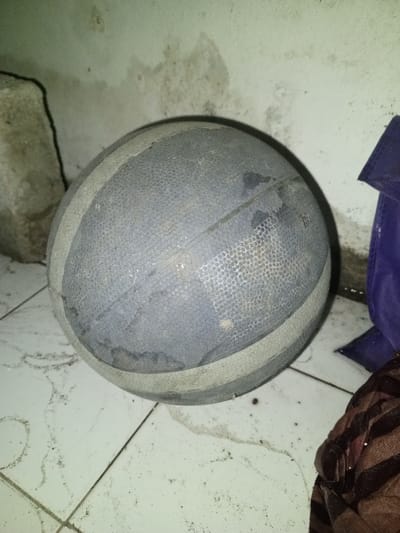 A used, dark grey basketball with two lighter grey circumferential stripes rests on a light-colored, multi-tiled floor. The basketball's surface exhibits significant wear, dust accumulation, and dark stains, with its pebbled texture visible but degraded in areas. The tiled floor beneath the ball is dirty, marked by numerous dark specks, smudges, and indistinct streaking patterns along the grout lines.
In the upper left background, a large, irregularly shaped, light-colored block, possibly concrete or stone, is positioned against a light-colored wall. This wall displays widespread dirt, dark splotches, and mildew-like stains. To the upper right, a section of dark blue fabric, potentially a bag or tarp, is visible. In the lower right, a crumpled piece of brown fabric with a dark, patterned design is present. The scene is dimly lit, suggesting an indoor or sheltered outdoor setting within Keo Kouang, Laos. No individuals are present, and no active interactions are depicted.