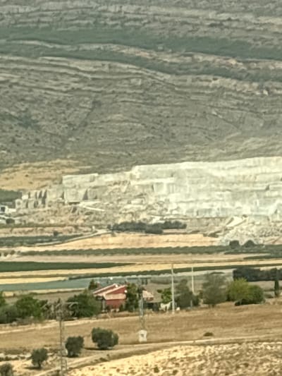 This is a long-range, aerial, daytime photograph showing a landscape in Caudete, Spain. The image is characterized by a hazy, desaturated color palette dominated by earth tones, greens, and whites. The foreground features dry, sandy terrain with sparse vegetation and a few trees. In the midground, a red-roofed building is visible, surrounded by greenery, and further back, agricultural fields stretch across the land. The background is dominated by a large, white, terraced hillside, indicative of a quarry or open-pit mine. The overall impression is one of a rural, arid environment with evidence of industrial activity.