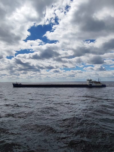 The image depicts a long, flat-bottomed barge floating on a body of water. The water is choppy with visible ripples and a dark gray hue. The barge is dark, likely a drab color, and extends nearly the width of the image, with a visible superstructure and details at each end. The sky is overcast with a mix of white and gray clouds and patches of blue. No people are visible on the boat or on the shore. It is unlikely that the location is the city of Mologa in Russia because that city does not currently exist; it was flooded in the 1940s to create the Rybinsk Reservoir. The image likely depicts a similar body of water as the Rybinsk Reservoir.