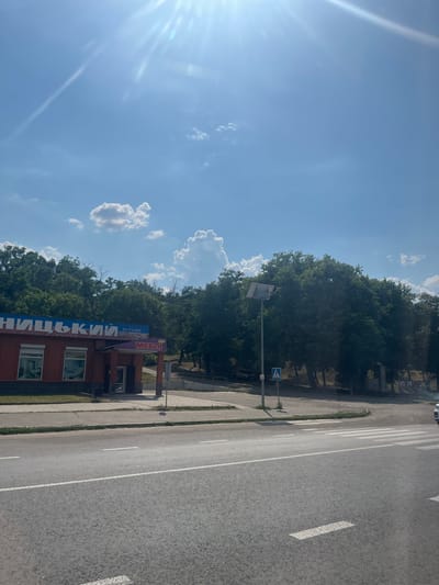 The image, taken in Balakleia, Ukraine, depicts a street scene under a bright, sunny sky with scattered clouds. In the foreground, a paved road with white lane markings and a pedestrian crossing is visible. To the left, a red brick building with large windows and a blue awning displaying Ukrainian text that translates to "FURNITURE STORE" and "MEBEL" (furniture) stands. Behind the building and extending across the middle ground is a dense line of green trees. A lamppost with a solar panel is situated near the building, next to a pedestrian crossing sign. The background shows more trees and some distant structures. The overall impression is of a commercial area on a clear day.