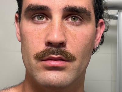 A close-up portrait of a young man with brown hair, a mustache, and green eyes. He is wearing two small silver hoop earrings in his right ear and a small silver ring in his left nostril. He is looking directly at the camera. The background is a white tiled wall, possibly in a bathroom.  The coordinates provided are for the city of Alon HaGalil, Israel. It is unclear if the image was taken in Alon HaGalil.