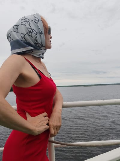 A woman with reddish hair, wearing a red ribbed dress and a patterned headscarf, stands on the deck of a vessel. She is wearing sunglasses and looking out at a body of water. Her left hand rests on her hip, and her right hand is on the railing. The water is dark and choppy, with a distant shoreline visible under a cloudy sky. The setting appears to be a wide river or lake, possibly near Bereg, Russia, given the context. The railing is white, and a rope runs along it. The overall scene suggests a journey or a moment of contemplation outdoors.