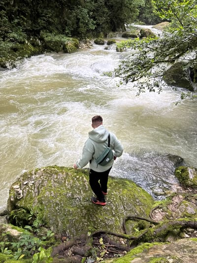 A person stands on a moss-covered rock outcropping, viewing a turbulent, milky-colored river flowing through a rocky gorge. The person wears a light-colored hooded sweatshirt, dark pants, and orange and black running shoes, with a small green bag slung across their body. Vegetation clings to the rocks on either side of the river, including a tree branch with leaves hanging into the scene from the upper left. The image is taken from above, creating a slightly distorted perspective. The location is Tsabal, Abkhazia.
