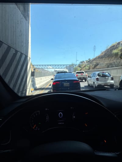 The image, taken from inside a vehicle, shows a traffic jam on a highway. Several vehicles are visible ahead, including a blue sedan and a white SUV. The background shows a hillside with vegetation under a clear sky. The dashboard of the vehicle in which the photo was taken is partially visible. The vehicle's odometer reads 170357 km, and the speedometer indicates 0 km/h. The "RUN" indicator is lit on the dashboard. The scene is a highway likely in the city of Atizapán de Zaragoza, Mexico.