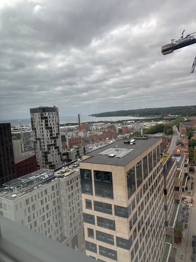 The image depicts an aerial view of the Aarhus Ø district in Aarhus, Denmark, under a cloudy sky. In the foreground, a large, modern building with a beige brick facade and dark-framed windows dominates the right side of the frame. Adjacent to it, to the left, is another contemporary building with a white, grid-like exterior. Further left, a tall, slender skyscraper with a patterned facade stands prominently. In the distance, a body of water is visible, likely the Aarhus Bay, with ships and industrial structures lining the waterfront. A construction crane, bearing the text "KRAN ELEKTRO", is visible in the upper right corner, indicating ongoing development in the area. The streets are lined with parked cars and bicycles, and some greenery is integrated into the urban landscape, with rooftop gardens visible on some buildings. The overall scene is one of a modern, developing urban environment with a clear presence of both residential and industrial architecture.
