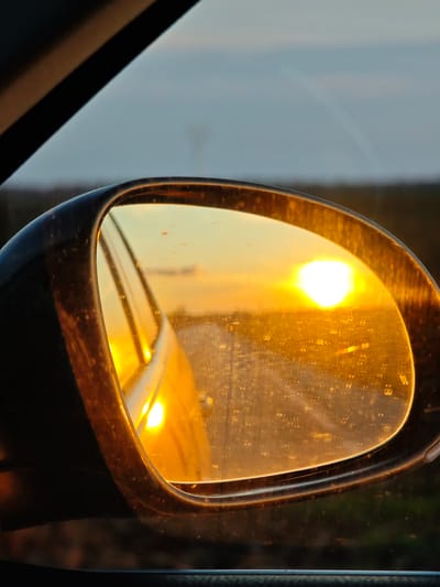 The image is a view of a sunset reflected in a car's side mirror. The mirror frame is dark and speckled with what appears to be dust or water spots. The sunset is bright orange, dominating the reflection with a bright glow in the center. A road stretches out towards the horizon in the mirror's reflection. Outside the mirror, a portion of the car's interior is visible, along with a glimpse of the sky and horizon. The exact location of the car is not discernible from the image. The location being Vaglen, Bulgaria, could not be determined.